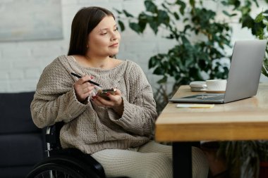 Tekerlekli sandalyede oturan genç bir kadın elinde kalem ve telefonuyla düşünceli bir şekilde oturuyor..