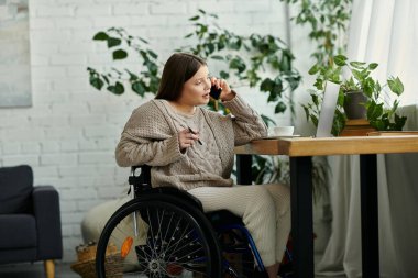 Tekerlekli sandalyedeki genç bir kadın evinde bir masada oturuyor ve telefonuyla konuşuyor..