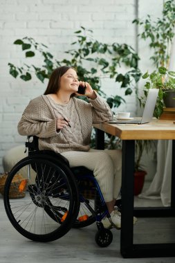 Tekerlekli sandalyedeki genç bir kadın masada oturuyor, telefonla konuşurken gülümsüyor. O, rahat ve aydınlık bir ortamda evde..