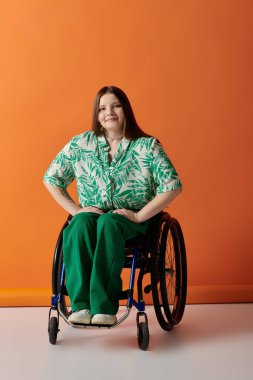 A young woman in a green and white floral shirt sits in a wheelchair against a bright orange background, smiling confidently at the camera. clipart