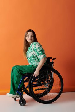Parlak yeşil kıyafetli kendine güvenen genç bir kadın parlak turuncu bir arka plana karşı tekerlekli sandalyede oturuyor ve gülümsüyor..