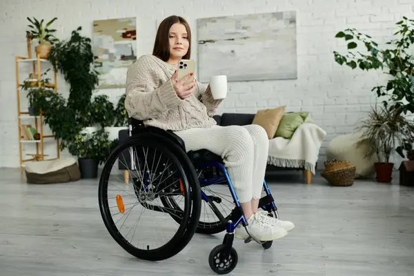 stock image A young woman sits in her wheelchair at home, using her phone and enjoying a cup of coffee.