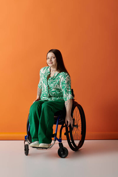 A confident young woman with a disability sits in a wheelchair, wearing green pants and a floral shirt against an orange background.