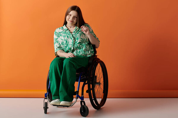 A woman with long brown hair, wearing colorful floral shirt and green pants, sits confidently in a wheelchair against orange background.