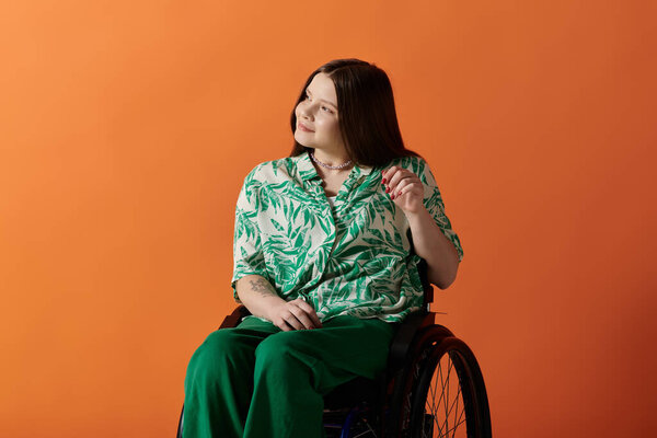 A smiling woman with long brown hair sits in a wheelchair against an orange backdrop, wearing green and white floral shirt and pants.