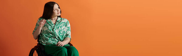 A young woman in a vibrant green and white patterned shirt sits in a wheelchair against a bright orange background.