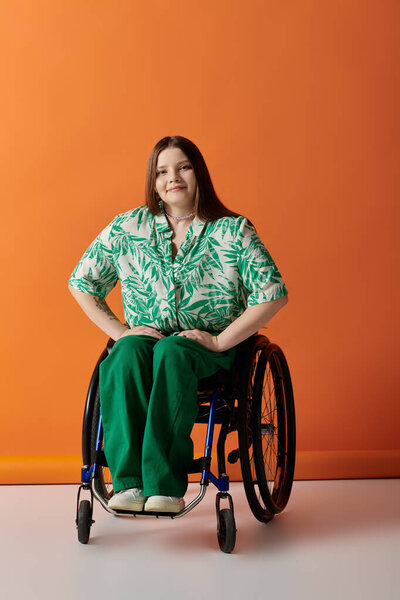A young woman in a green and white floral shirt sits in a wheelchair against a bright orange background, smiling confidently at the camera.