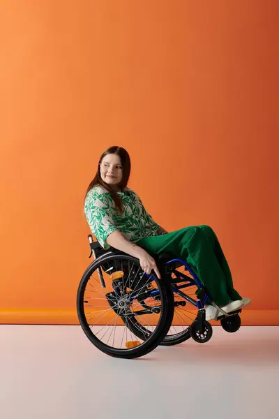 stock image A young woman in vibrant attire sits confidently in a wheelchair against a bright orange background.