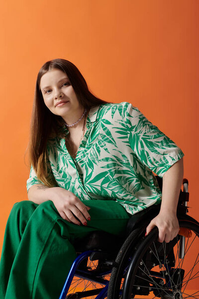 A young woman wearing green clothing with a leafy pattern smiles confidently while sitting in a wheelchair against an orange background.