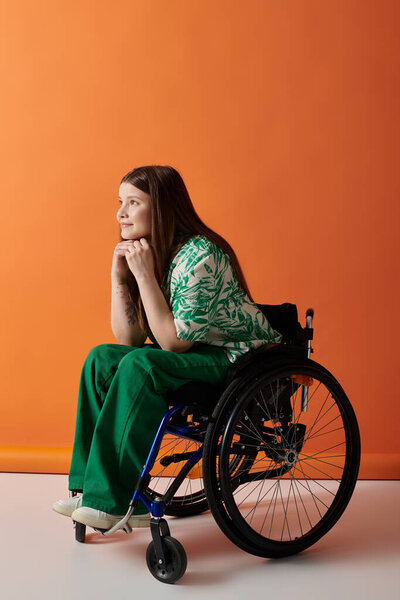 A young woman in a vibrant green outfit sits in a wheelchair against an orange background, looking thoughtfully ahead.