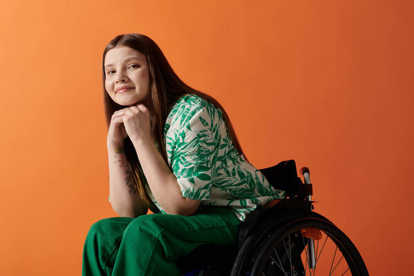 A disabled young woman in a green dress sits gracefully in a wheelchair against an orange backdrop.