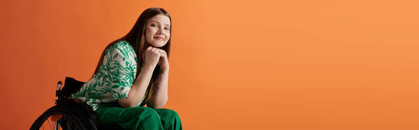 A disabled young woman in a vibrant green dress sitting confidently in a wheelchair against an orange background.