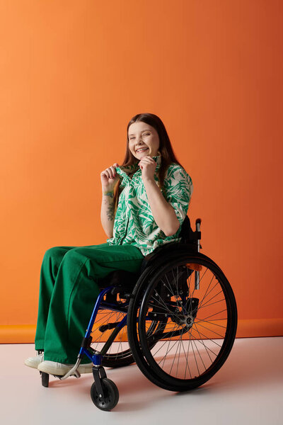 A young woman in a vibrant green shirt and pants sits in a wheelchair against an orange background, smiling.