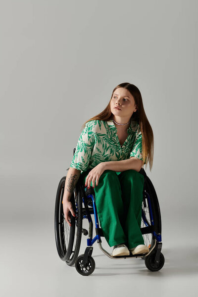 Молодая женщина в зеленом сидит в инвалидном кресле на сером фоне.