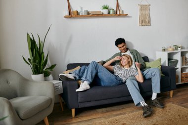 A young multicultural couple laughs and enjoys each others company on a comfortable couch in their modern apartment. clipart