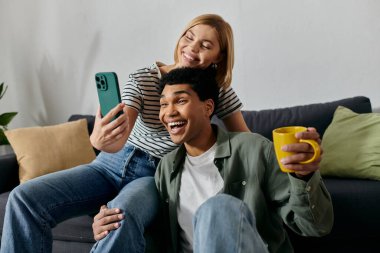 Genç, çok kültürlü bir çift modern dairelerinde telefonla selfie çekerken gülümsüyor ve gülüyor..