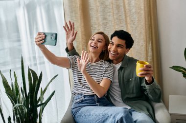 Genç, çok kültürlü bir çift modern dairelerinde selfie çekmek için gülümsüyor, rahat ve samimi bir an sergiliyor..