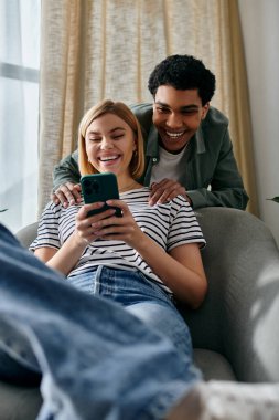 Genç, çok kültürlü bir çift evlerindeki modern bir kanepede rahatlar, akıllı telefona bakarken birlikte gülerler..