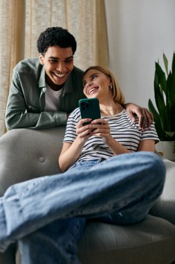Genç, çok kültürlü bir çift modern bir dairede koltukta oturup gülüyor ve akıllı telefona bakıyor..
