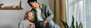 A young multicultural couple shares a laugh while spending time in their modern apartment. clipart