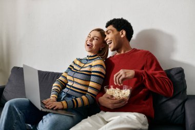 Mutlu bir ırklar arası çift, rahat, modern dairelerinde dizüstü bilgisayarlarında film izlerken birlikte güler..