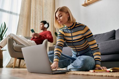 A young woman uses a laptop while her partner relaxes in the background, enjoying their time together in a modern apartment. clipart