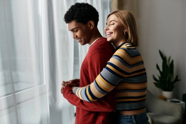 Mutlu, çok kültürlü bir çift modern bir apartmanın penceresinin önünde kucaklaşır..