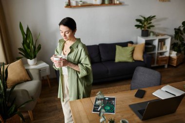 A woman stands by her desk, taking a break from work with a cup of coffee in her hand. clipart