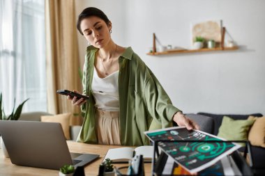Bir kadın ofis masasında duruyor, telefonunu tutuyor ve kağıtları düzenliyor..