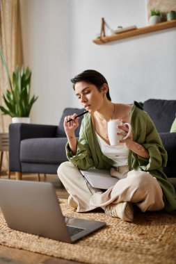 Bir kadın dizüstü bilgisayar ve bir fincan kahveyle yerde oturuyor, düşüncelere dalmış..