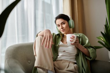 Yeşil gömlekli bir kadın evde oturup kulaklıkla müzik dinliyor ve kahve içiyor..