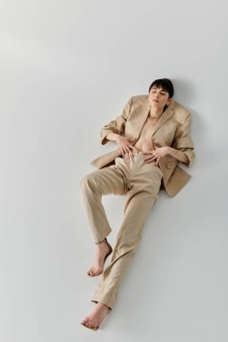 Bej takım elbiseli kadın beyaz arka planda uzanmış, bacakları açık, elleri belinde.