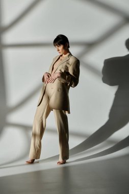 Woman in tan suit poses in front of white background, sunlight casting shadows. clipart