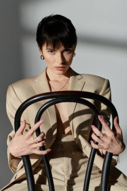 A woman in a beige suit poses while sitting on a black chair. clipart