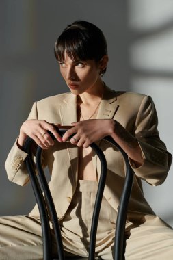 Bej ceketli bir kadın stüdyo ortamında siyah bir sandalyeye yaslanıyor..