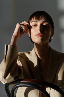 A woman with short, dark hair poses in a light-filled room, sitting on a chair. clipart