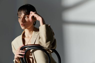 Woman in tan blazer sits in chair, posing thoughtfully amid light and shadows. clipart