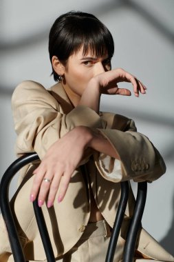 A woman wearing a tan blazer poses in a studio with a sunlit backdrop. clipart