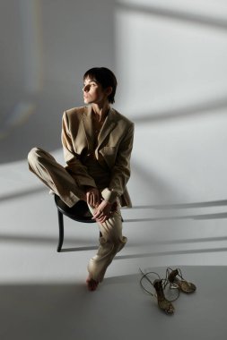 Bronz takım elbiseli bir kadın beyaz bir arka plana karşı bir taburede oturuyor, düşüncelere dalmış..