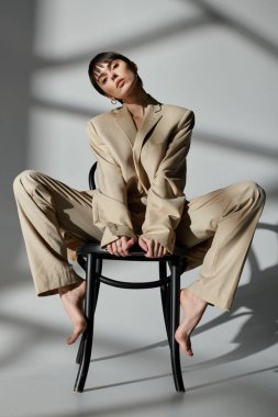 A woman in a tan suit sits on a black chair in a studio setting. clipart