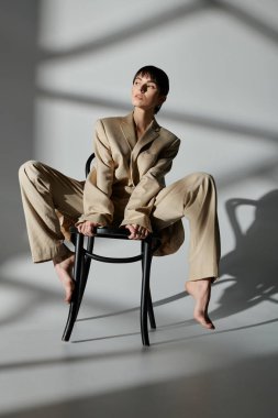 A woman in a beige suit poses on a chair against a white backdrop. clipart