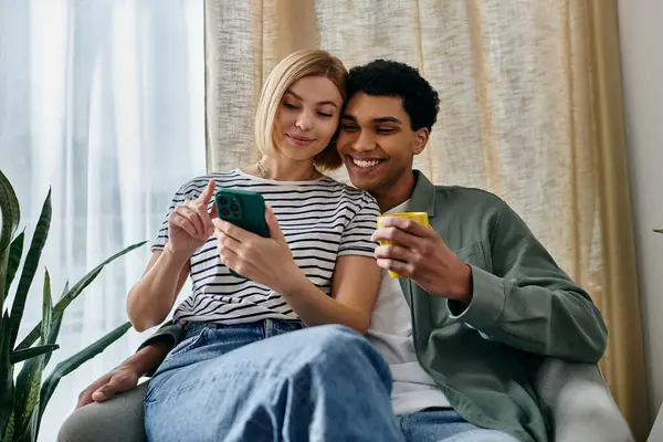 stock image A young multicultural couple enjoys a relaxing afternoon in their modern apartment, looking at a phone and sharing a cup of tea.