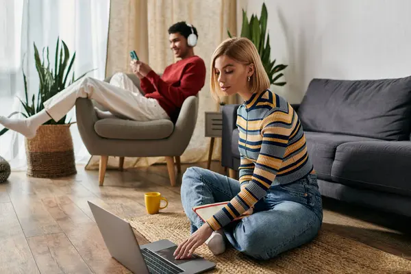 stock image A happy couple relaxes in their modern apartment, one woman on the floor using a laptop while the other man sits in a chair.