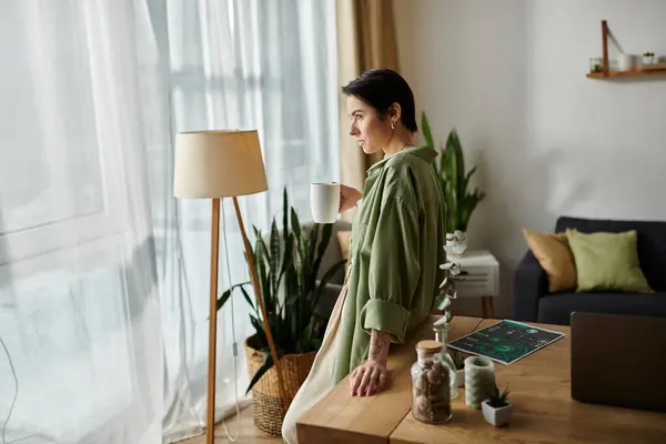 stock image A woman stands at a desk in her home office, holding a cup of coffee and looking out the window.