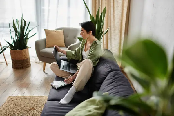 stock image A woman sits on a couch, working on a laptop, while surrounded by houseplants.