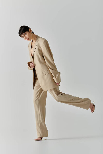 A young woman in a tan suit stands against a white background, posing with one leg extended.
