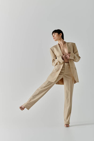 A woman in a tan suit stands in a white studio, one leg extended forward.