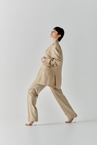 A woman in a stylish beige suit walks confidently on a white backdrop.