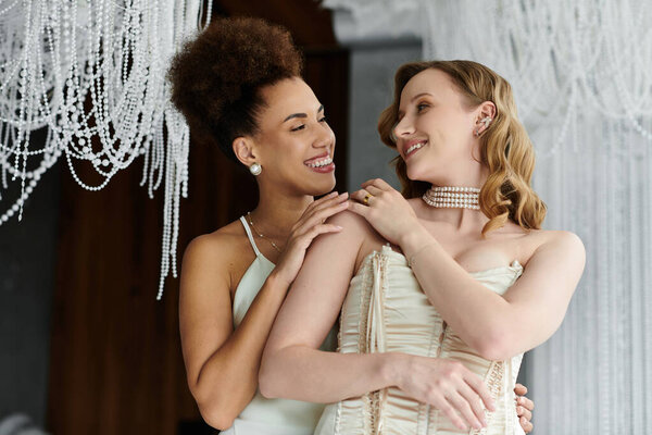 Две будущие невесты обнимаются под сверкающей люстрой, их счастье излучается.