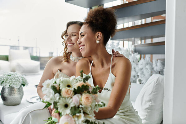 Two brides smiling tenderly, gazing out a window on their wedding day.